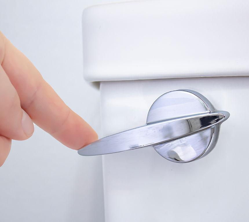 A close-up of a hand about to push down a silver flush lever on a white toilet tank.